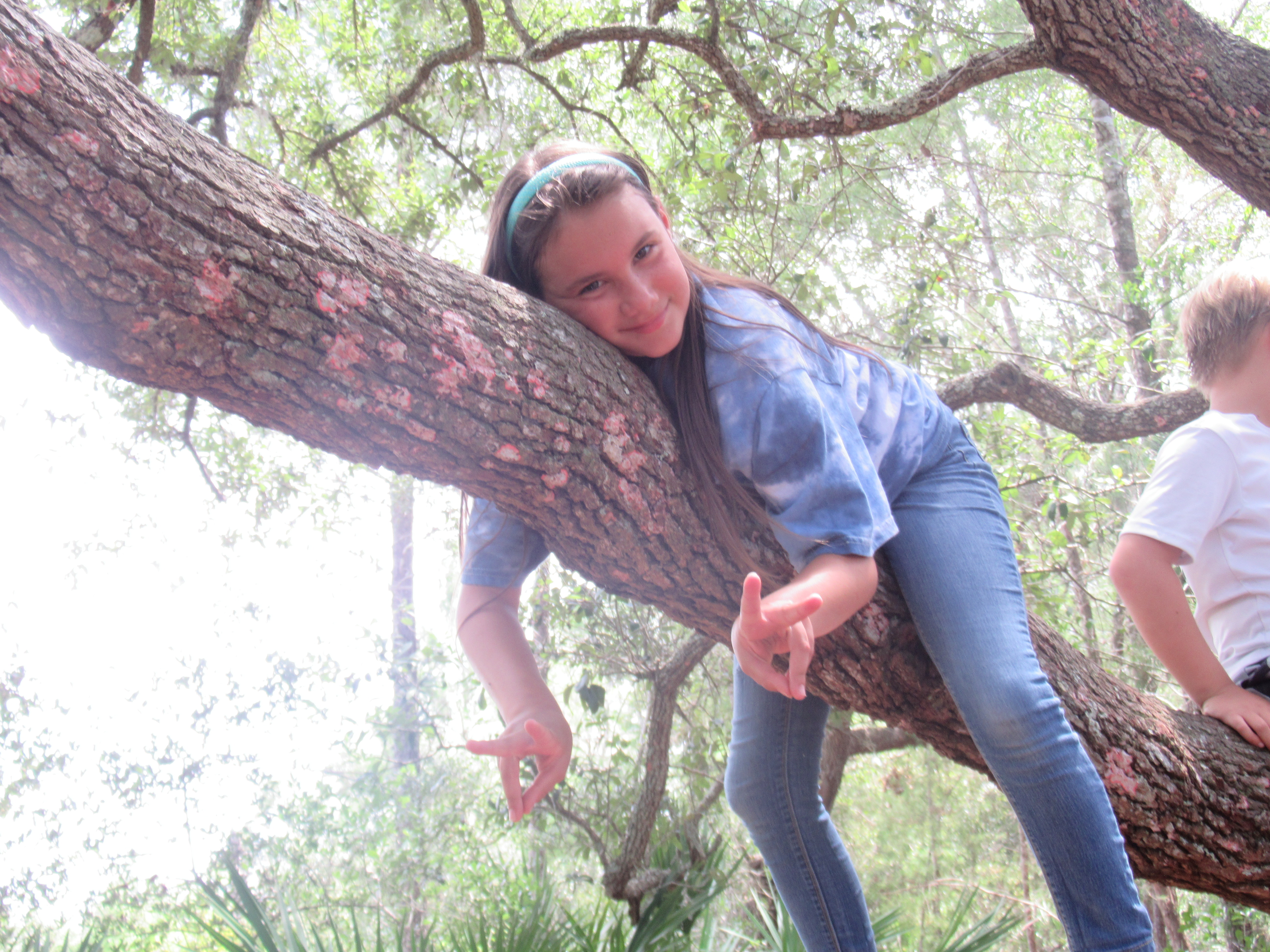 Girl laying on tree branch, smiling, giving peace signs with her hands